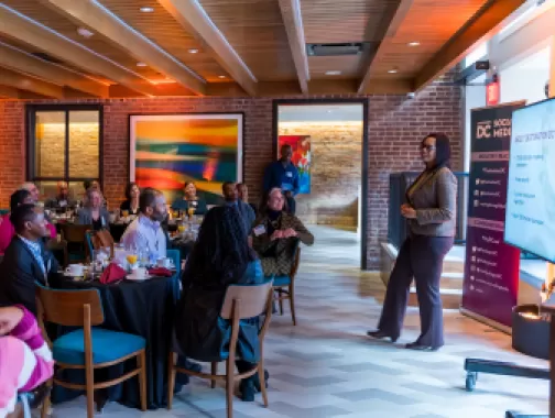 A woman presents information about Destination DC to an engaged audience seated at round tables during a professional breakfast event in a warmly lit venue.
